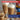 Two tall clear glass mugs filled with a layered coffee drink topped with whipped cream and a sprinkle of cinnamon, shown on a wooden surface with a bowl of nuts and a Torani syrup bottle in the background. Promotional text overlays read '33% More Flavor, for less.